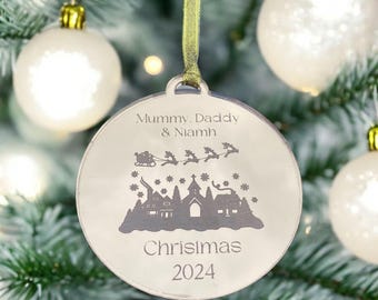 May include: A silver round Christmas ornament with a white background and a black silhouette of a snowy village scene with Santa Claus flying over the village. The ornament is personalized with the text "Mummy, Daddy & Niamh Christmas 2024".