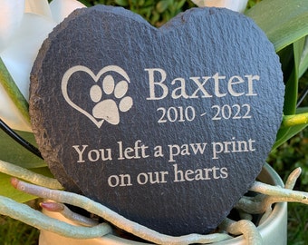 May include: A black heart-shaped slate stone memorial with the inscription "Baxter 2010 - 2022 You left a paw print on our hearts" and a paw print inside a heart.