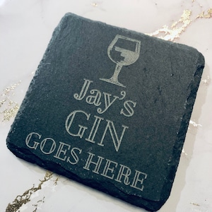 May include: Square slate coaster with etched text and a wine glass graphic. The text reads "Jay's GIN GOES HERE". The coaster is dark gray and has a rough, natural edge. A perfect gift for gin lovers.