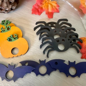 May include: A set of Halloween-themed wooden decorations, featuring pumpkins, bats and spiders. The pumpkins are orange with green stems, the bats are dark blue and the spiders are black. All of the decorations have a hole in the centre for hanging.