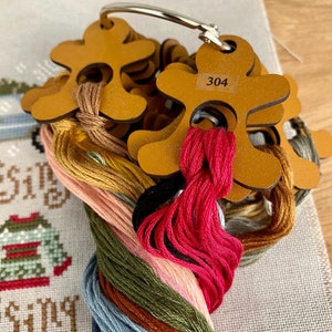 May include: A set of colourful embroidery floss wound around wooden thread organisers shaped like gingerbread people. The organisers are numbered, with one labelled "304".