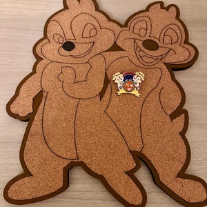 Puede incluir: Un tablero de corcho con forma de dos ardillas de dibujos animados, una marrón y otra beige, de pie una al lado de la otra. Las ardillas sonríen y tienen ojos negros. Un pequeño alfiler con una imagen de dibujos animados de dos ardillas está sujeto al tablero de corcho.