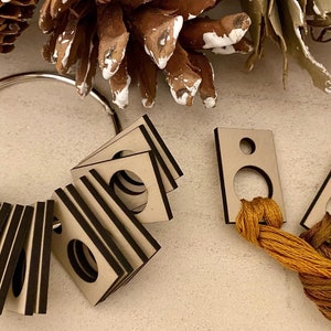 May include: A set of wooden thread organisers with circular holes, stacked on a metal ring. The organisers are brown with black edges. A single organiser is shown separately with a length of orange embroidery floss threaded through the hole.