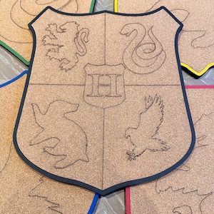 Può includere: Scudo in sughero con bordo nero, con disegni incisi di un leone, un serpente, un tasso e un'aquila, con una "H" al centro. Sono visibili altri scudi con bordi colorati, tra cui verde, giallo, blu e rosso.