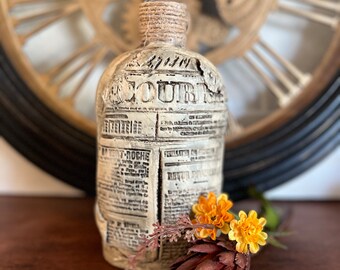 May include: A decorative bottle with a newspaper-like design and the word "COURT" printed on it. The bottle is wrapped with twine at the top and is sitting on a wooden surface. There are dried flowers next to the bottle.