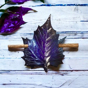 Puede incluir: Horquilla artesanal con forma de hoja de arce. La hoja es un degradado de morado y negro, con un palito de madera para sujetar el cabello. La horquilla está sobre un fondo de madera blanca, con hojas moradas en la esquina superior izquierda. Mide unos 10 cm.