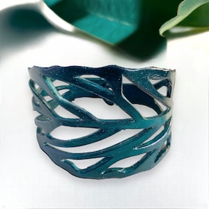 May include: A teal cuff bracelet with a cut-out leaf design. The bracelet is made of a flexible material and has a textured surface. The openwork design allows light to pass through, creating a unique visual effect.