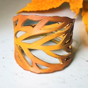 May include: A wide, open-work cuff bracelet in shades of brown and yellow. The leather bracelet features a leaf-like design with cut-out shapes. The bracelet is approximately 5 cm wide.