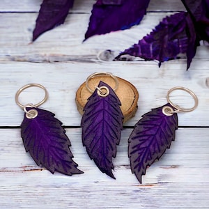 Puede incluir: Tres llaveros de cuero morado con forma de hoja con anillas plateadas. Los llaveros están colocados sobre una superficie de madera clara, con una rodaja de madera en el fondo. Las hojas tienen vetas detalladas y un color morado oscuro.