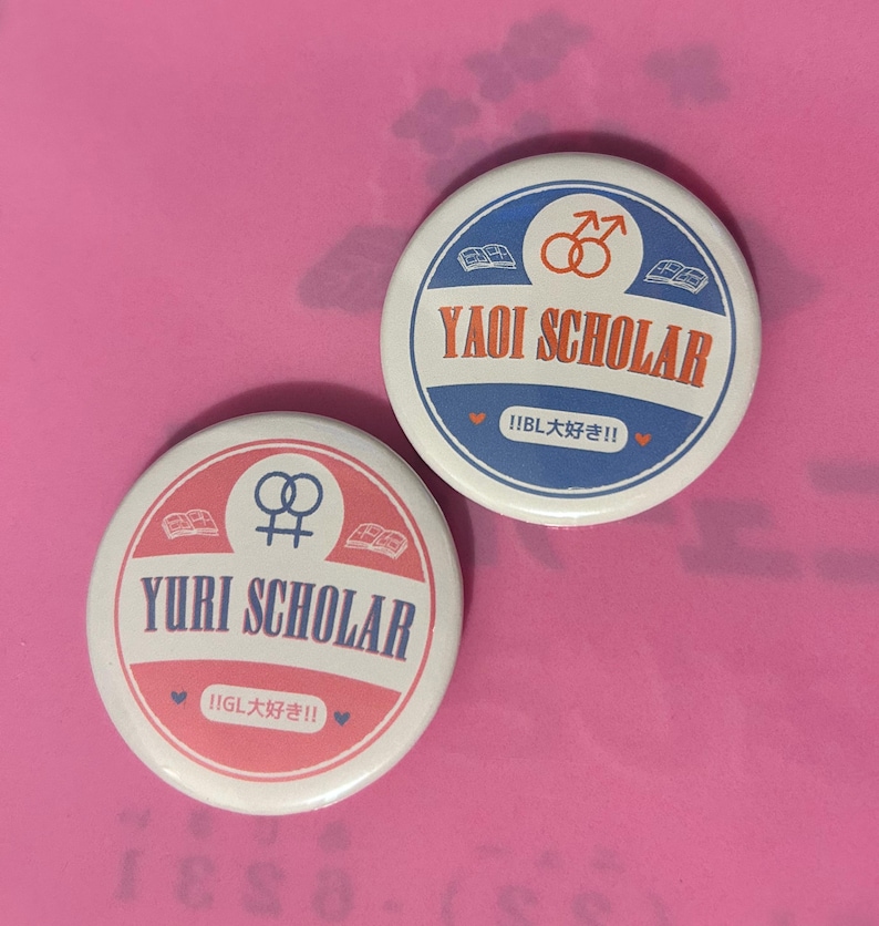 May include: Two round buttons with text and symbols on a pink background. One button reads "YURI SCHOLAR" in white text on a pink and white background. The other button reads "YAOI SCHOLAR" in white text on a blue and white background.