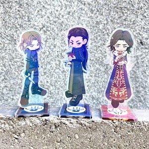 Könnte beinhalten: Drei Acryl-Charakter-Standees mit Illustrationen im Anime-Stil. Die Figuren stellen Charaktere in verschiedenen Outfits dar, die auf transparenten Sockeln stehen. Die Standees haben detaillierte Designs und lebendige Farben.