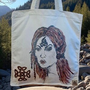 Può includere: Una borsa tote bianca con un'illustrazione disegnata a mano di una donna con i capelli rossi e disegni di nodi celtici. La borsa è appesa a un ramo d'albero sullo sfondo di montagne e un cielo soleggiato.
