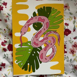 May include: A colorful art print featuring a pink snake with dark pink spots, a forked tongue, and a green leaf design. The background is yellow with white wavy shapes. The art print is on a white background.