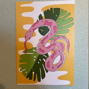 May include: A pink snake illustration with dark pink spots and a red tongue, set against a yellow and white background with green tropical leaves. The artwork has a playful, cartoonish style.
