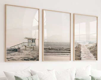 May include: Three framed photographs of a beach scene with a lifeguard stand, a wooden walkway, and a view of the ocean. The photographs are hung on a white wall above a white couch with pillows and a throw blanket.