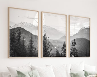 May include: Three black and white framed photographs of a mountain range with trees in the foreground. The photographs are hanging on a white wall above a white couch with pillows and a throw blanket.