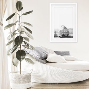 May include: A white wicker daybed with a white throw blanket and grey pillows. A framed black and white photograph of a vintage camper van is hanging on the wall above the daybed. A large green plant in a white wicker basket is in the foreground.