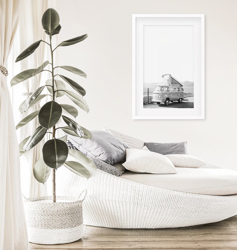 May include: A white wicker daybed with a white throw blanket and grey pillows. A framed black and white photograph of a vintage camper van is hanging on the wall above the daybed. A large green plant in a white wicker basket is in the foreground.
