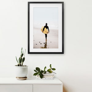 Puede incluir: Una fotograf&iacute;a enmarcada en negro de una persona caminando por una playa con una tabla de surf bajo el brazo. La playa es arenosa y el agua es poco profunda. El cielo est&aacute; nublado y el fondo est&aacute; desenfocado.