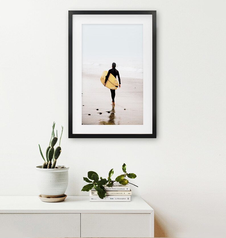 Puede incluir: Una fotograf&iacute;a enmarcada en negro de una persona caminando por una playa con una tabla de surf bajo el brazo. La playa es arenosa y el agua es poco profunda. El cielo est&aacute; nublado y el fondo est&aacute; desenfocado.