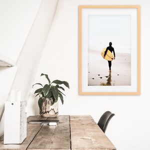 Puede incluir: Una fotograf&iacute;a enmarcada de una persona con un traje de neopreno caminando por una playa con una tabla de surf bajo el brazo. La foto est&aacute; en un marco de madera clara y est&aacute; colgada en una pared blanca.