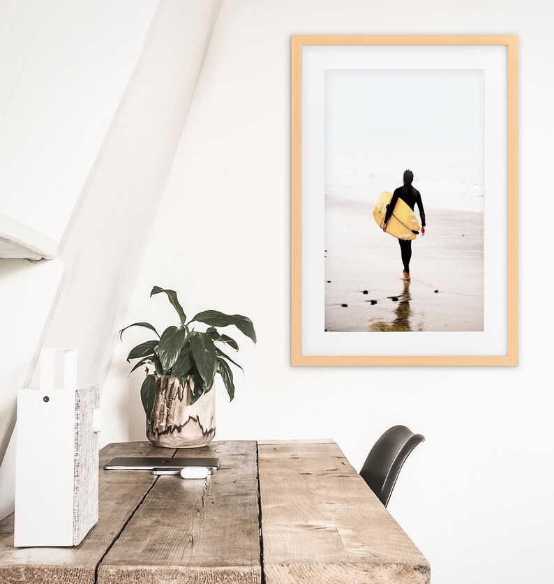 Puede incluir: Una fotograf&iacute;a enmarcada de una persona con un traje de neopreno caminando por una playa con una tabla de surf bajo el brazo. La foto est&aacute; en un marco de madera clara y est&aacute; colgada en una pared blanca.