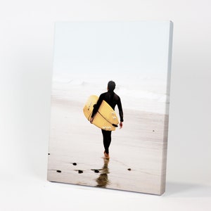 Puede incluir: Una persona con un traje de neopreno negro camina por una playa de arena llevando una tabla de surf amarilla.