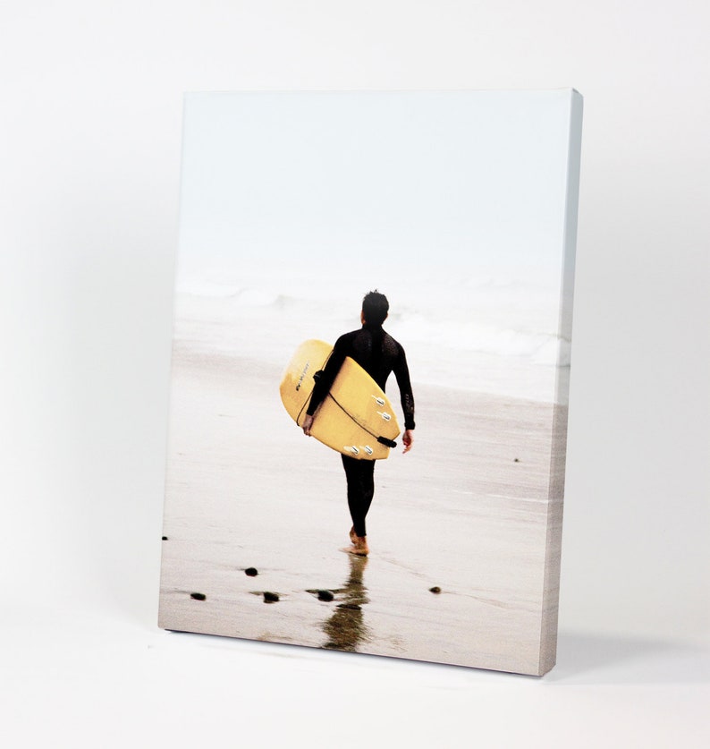 Puede incluir: Una persona con un traje de neopreno negro camina por una playa de arena llevando una tabla de surf amarilla.