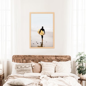 Puede incluir: Una fotograf&iacute;a enmarcada de una persona caminando por una playa con una tabla de surf. La persona lleva un traje de neopreno y lleva la tabla de surf sobre el hombro. La playa es arenosa y el agua es azul.