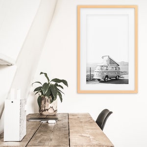 May include: A black and white photograph of a vintage camper van parked on a dirt road. The van has a pop-up roof and is surrounded by mountains. The photo is framed in a light wood frame.