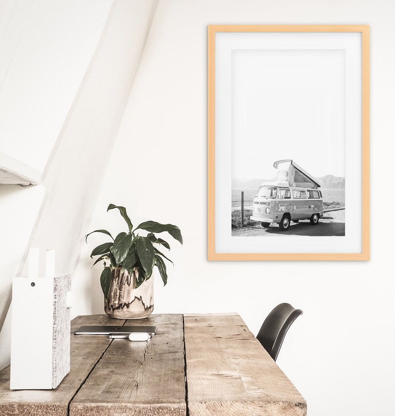 May include: A black and white photograph of a vintage camper van parked on a dirt road. The van has a pop-up roof and is surrounded by mountains. The photo is framed in a light wood frame.