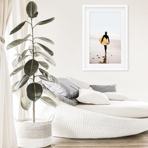 Puede incluir: Un div&aacute;n de mimbre blanco con almohadas de estampado gris y blanco. Una gran planta verde est&aacute; en una cesta de mimbre blanca en primer plano. Una fotograf&iacute;a enmarcada de una persona caminando por la playa con una tabla de surf est&aacute; en la pared.