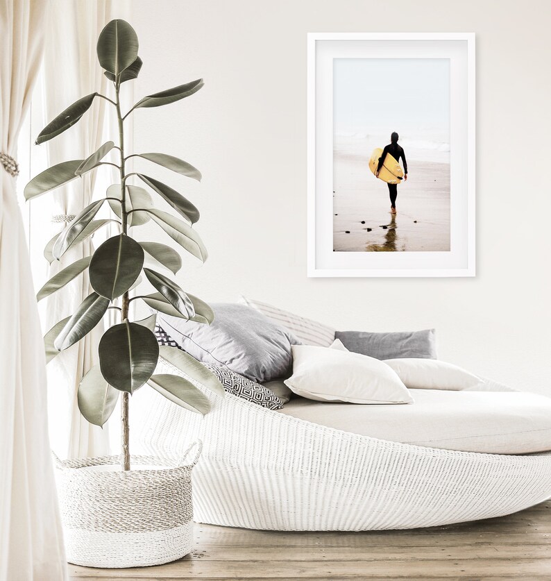 Puede incluir: Un div&aacute;n de mimbre blanco con almohadas de estampado gris y blanco. Una gran planta verde est&aacute; en una cesta de mimbre blanca en primer plano. Una fotograf&iacute;a enmarcada de una persona caminando por la playa con una tabla de surf est&aacute; en la pared.