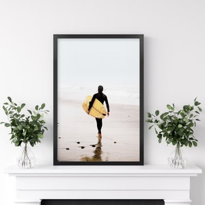 Puede incluir: Una fotograf&iacute;a enmarcada en negro de una persona caminando por una playa con una tabla de surf amarilla. La persona lleva un traje de neopreno negro y camina de espaldas a la c&aacute;mara. La playa es arenosa y el agua es clara.