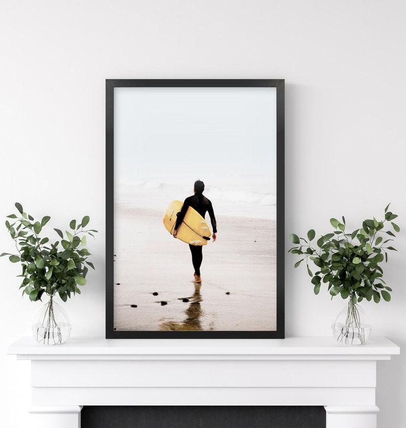 Puede incluir: Una fotograf&iacute;a enmarcada en negro de una persona caminando por una playa con una tabla de surf amarilla. La persona lleva un traje de neopreno negro y camina de espaldas a la c&aacute;mara. La playa es arenosa y el agua es clara.