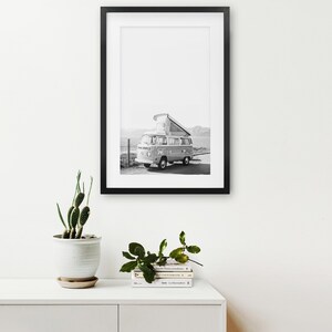 May include: Black and white photograph of a vintage camper van parked on a beach. The van has a pop-up roof and is facing the sea. The photo is framed in a black frame.