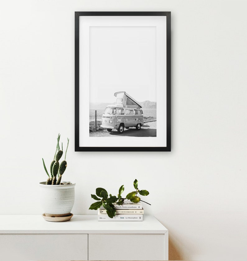 May include: Black and white photograph of a vintage camper van parked on a beach. The van has a pop-up roof and is facing the sea. The photo is framed in a black frame.