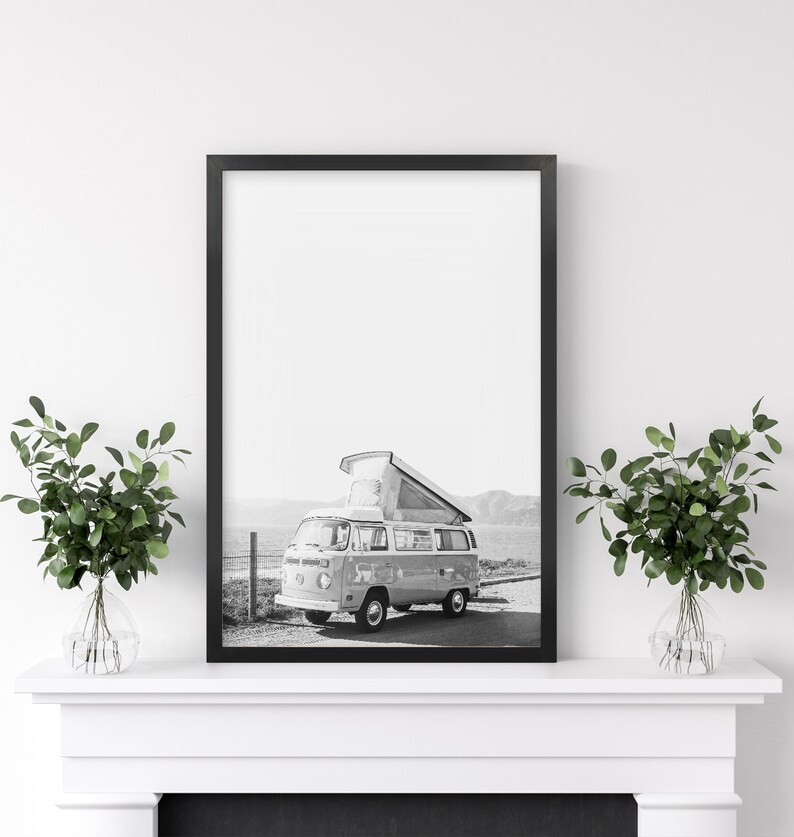 May include: Black and white photograph of a vintage camper van with a pop-up roof parked in a field. The van is facing the camera and the photo is framed in a black frame.