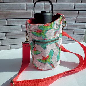 May include: A pink fabric water bottle holder with a parrot print and a red adjustable strap. The holder has a zippered pocket and a black water bottle inside.