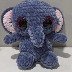 May include: A handmade blue crochet elephant plush toy with large, dark eyes and pink inner ears. The elephant has a trunk, small arms, and legs. The plush toy is made of soft, textured yarn.