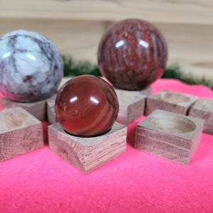 May include: Three polished gemstone spheres displayed on wooden stands. The spheres are red, white, and gray with intricate patterns. The stands are made of natural wood and have a rustic finish.