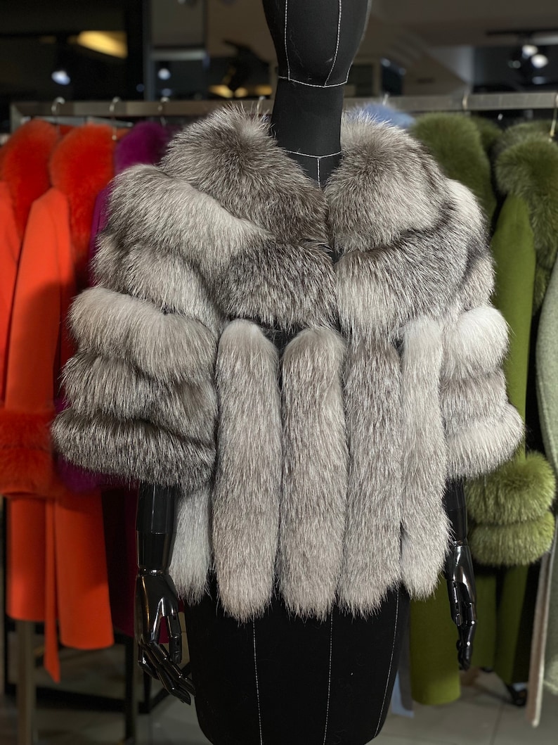 Puede incluir: Un chal de piel de zorro plateado con un cuello ancho y mangas largas. El chal se muestra en un maniqu&iacute; negro en una tienda.
