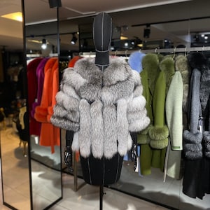 Puede incluir: Una estola de piel de zorro plateado con un dise&ntilde;o largo y fluido. La estola se exhibe en un maniqu&iacute; negro en un entorno de tienda minorista.