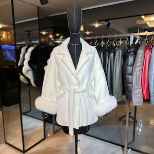 Op de afbeelding: Een witte nep bontjas met een riem en bontbeleg aan de manchetten. De jas is op een mannequin in een winkel.
