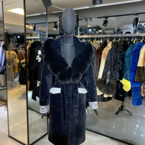 Könnte beinhalten: Ein schwarzer Pelzmantel mit einem großen schwarzen Pelzkragen, silbernen Verzierungen und einem Gürtel. Der Mantel befindet sich auf einer Schaufensterpuppe in einem Geschäft.