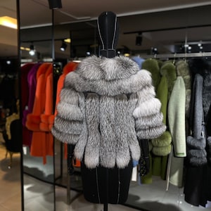 Puede incluir: Un abrigo de piel de zorro plateado de estilo corto y recortado. El abrigo se exhibe en un maniqu&iacute; negro en un entorno de tienda minorista.