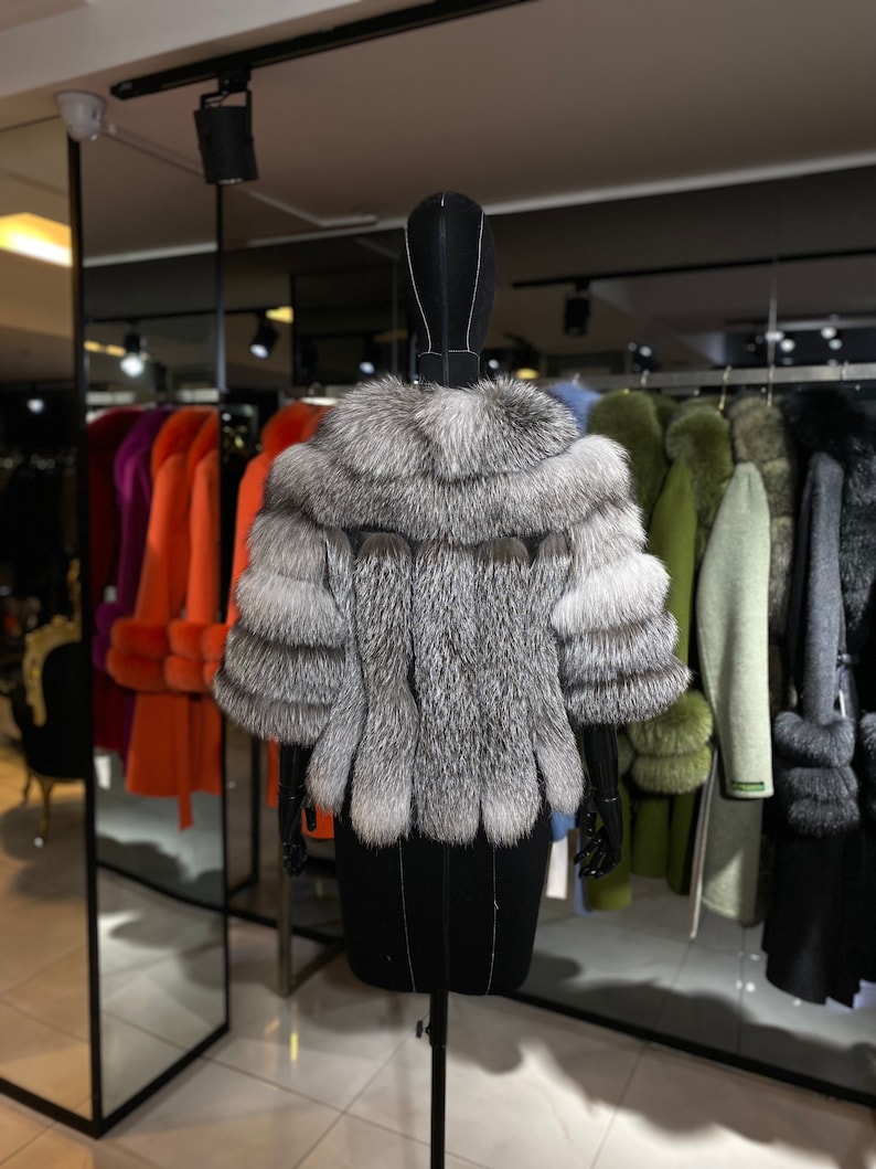 Puede incluir: Un abrigo de piel de zorro plateado de estilo corto y recortado. El abrigo se exhibe en un maniqu&iacute; negro en un entorno de tienda minorista.