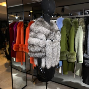 Puede incluir: Una chaqueta de piel de zorro plateado en un maniqu&iacute; negro en una tienda de ropa. La chaqueta tiene un estilo largo y fluido y est&aacute; hecha de piel suave y lujosa.