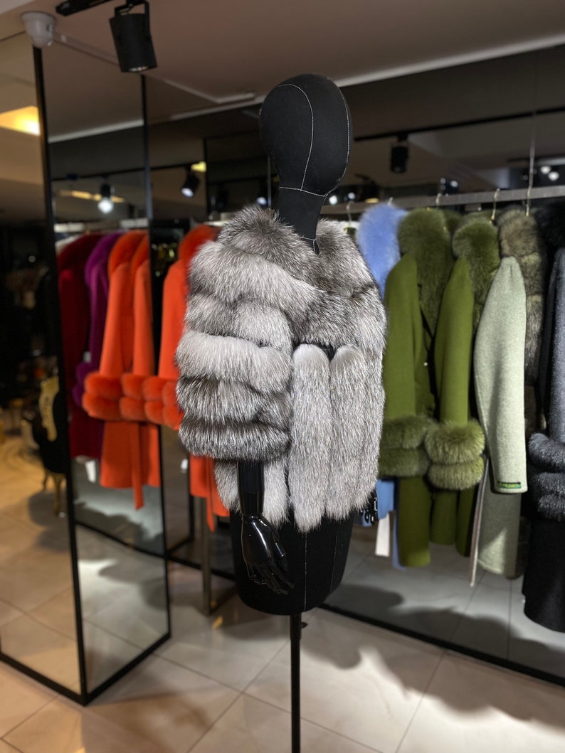 Puede incluir: Una chaqueta de piel de zorro plateado en un maniqu&iacute; negro en una tienda de ropa. La chaqueta tiene un estilo largo y fluido y est&aacute; hecha de piel suave y lujosa.