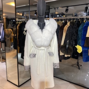 Könnte beinhalten: Ein weißer Pelzmantel mit einem großen weißen Pelzkragen und einem Gürtel. Der Mantel hat ein Schlangenhautmuster an den Taschen und Ärmelbündchen.